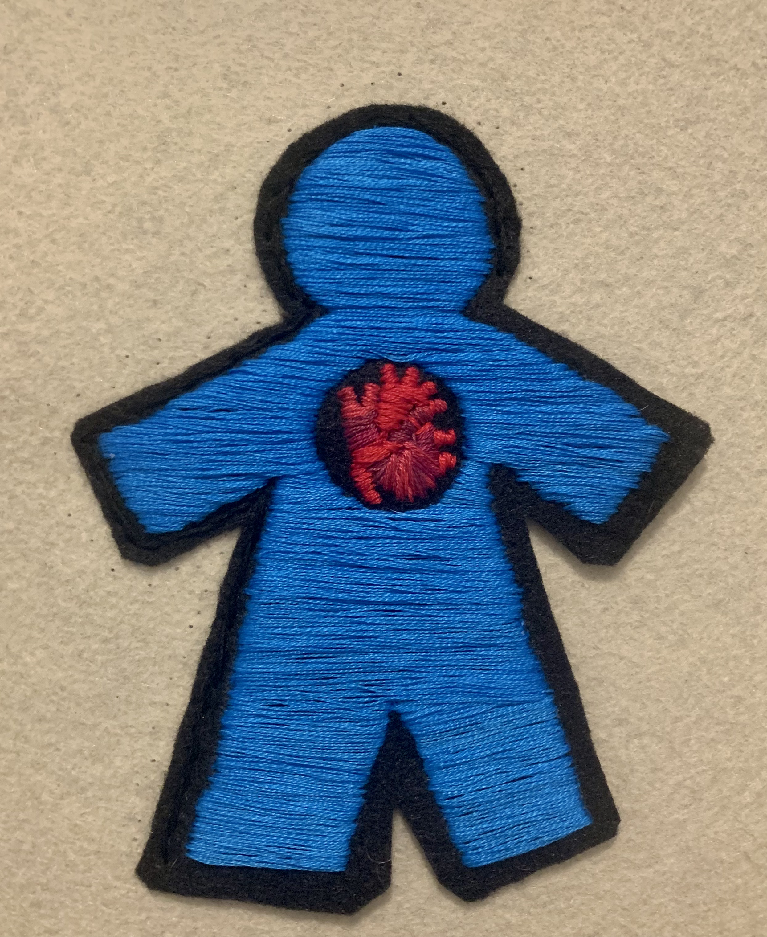 hand stitched felt patch, meant to look like a paper doll with a large hole in its chest. Inside the hole is a human heart. The doll’s body is blue and the heart isa mix of red, magenta, and pink. 